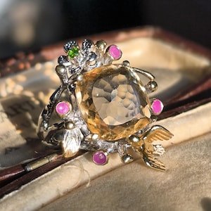 Op de afbeelding: Een gouden ring met een grote, gefacetteerde, gele edelsteen omringd door kleinere roze en groene edelstenen. De ring heeft een delicaat, bloemig ontwerp met een vogel die op de ene kant zit.