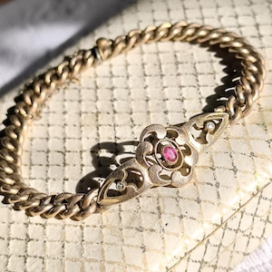 Könnte beinhalten: Ein goldfarbenes Armband mit einem Panzerketten-Design. Das Mittelstück zeigt ein Blumenmotiv mit einem zentralen roten Edelstein und kleinen, klaren Steinen. Das Armband wird auf einer strukturierten, cremefarbenen Oberfläche präsentiert.