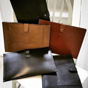 May include: A collection of leather document holders in shades of brown and black. Each has a flap closure with a button or clasp. Some have a front pocket. The items are displayed on a white chair.