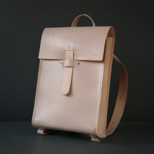 May include: A light beige leather backpack with a flap closure and adjustable straps. The bag has a top handle and a vertical strap closure with metal rivets. The bag is made of smooth, natural-toned leather.