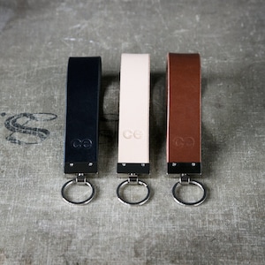 May include: Three leather keyrings in black, cream, and brown, each with a silver-tone ring and a metal accent. The keyrings have the initials "ce" embossed on them. The keyrings are displayed on a textured surface.