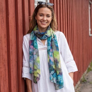 Peut inclure: Une robe longue blanche à manches longues avec des détails en dentelle au niveau de l'encolure et des manches, associée à une écharpe légère à motif floral abstrait multicolore dans des tons bleu, vert et violet, et un petit sac à main noir tressé.