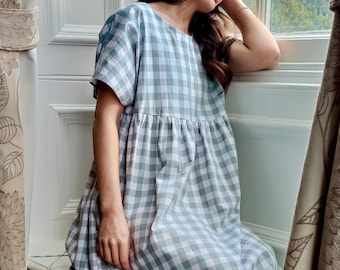 May include: A light blue and white gingham pattern dress with short sleeves. The dress has a loose, flowing silhouette. The image is taken indoors, near a window with floral curtains.