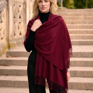 May include: A deep burgundy rectangular shawl featuring a soft, solid-colored fabric with delicate fringe detailing along the edges. The wrap is draped over the shoulders, showcasing its smooth texture and rich, dark red hue in a simple, classic design.
