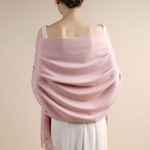 May include: A pale pink shawl draped over the shoulders. The shawl has a soft texture and a fringed edge. The person is wearing a white top and the background is a neutral colour.