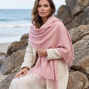 May include: A soft, solid light pink scarf with a smooth texture and fringed edges. The scarf is draped over the shoulders and upper body, featuring a rectangular shape with delicate tassels along the bottom hem, providing a simple, clean design.