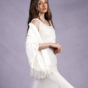 May include: A woman wearing a white wedding dress with a cowl neckline and a matching shawl with fringe. She is also wearing a silver tiara with star-shaped accents. The background is a solid lavender color.