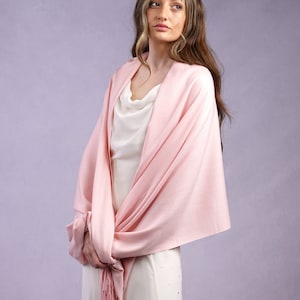 May include: A light pink shawl with a fringed edge is draped over a white dress. A silver tiara with star-shaped accents adorns the hair. The shawl is knotted at the front, creating a layered effect. The dress has a cowl neckline.