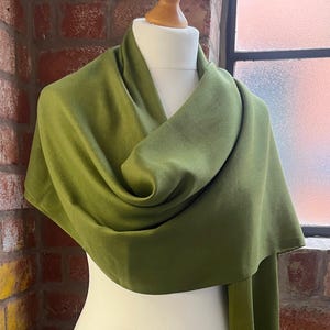 Op de afbeelding: Een olijfgroene sjaal gedrapeerd over een witte mannequin. De sjaal is gemaakt van een zachte stof en heeft een lang, vloeiend ontwerp. De achtergrond is een bakstenen muur en een raam.