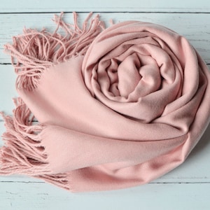 May include: A rolled-up, light pink scarf with fringed edges. The scarf is made of a soft material and is displayed on a white wooden surface. The scarf's color is a muted, dusty rose, and the fringe adds a textural element.