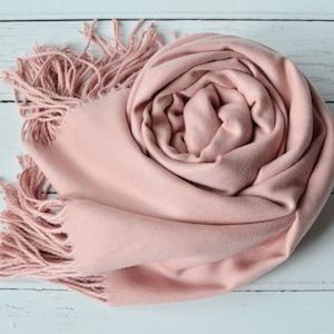 May include: A soft, blush pink scarf with a smooth, fine-knit texture and long, twisted fringe detailing at the ends. The scarf is loosely coiled into a circular shape, showcasing the consistent pastel pink color and the delicate fabric weave.