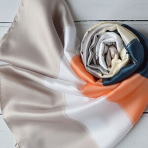 May include: A striped silk scarf in shades of beige, white, orange, and teal. The scarf is folded and rolled into a spiral shape.