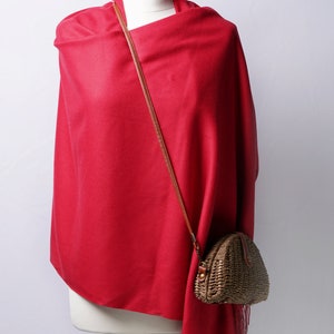 Peut inclure: Un châle rouge vif drapé sur un mannequin, accompagné d'un petit sac à main marron tissé avec une bandoulière en cuir. Le châle a une bordure à franges, et le sac est de forme triangulaire arrondie.