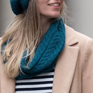 May include: A woman wearing a teal blue knitted beret and a matching teal blue infinity scarf. She is wearing a beige coat over a striped shirt.