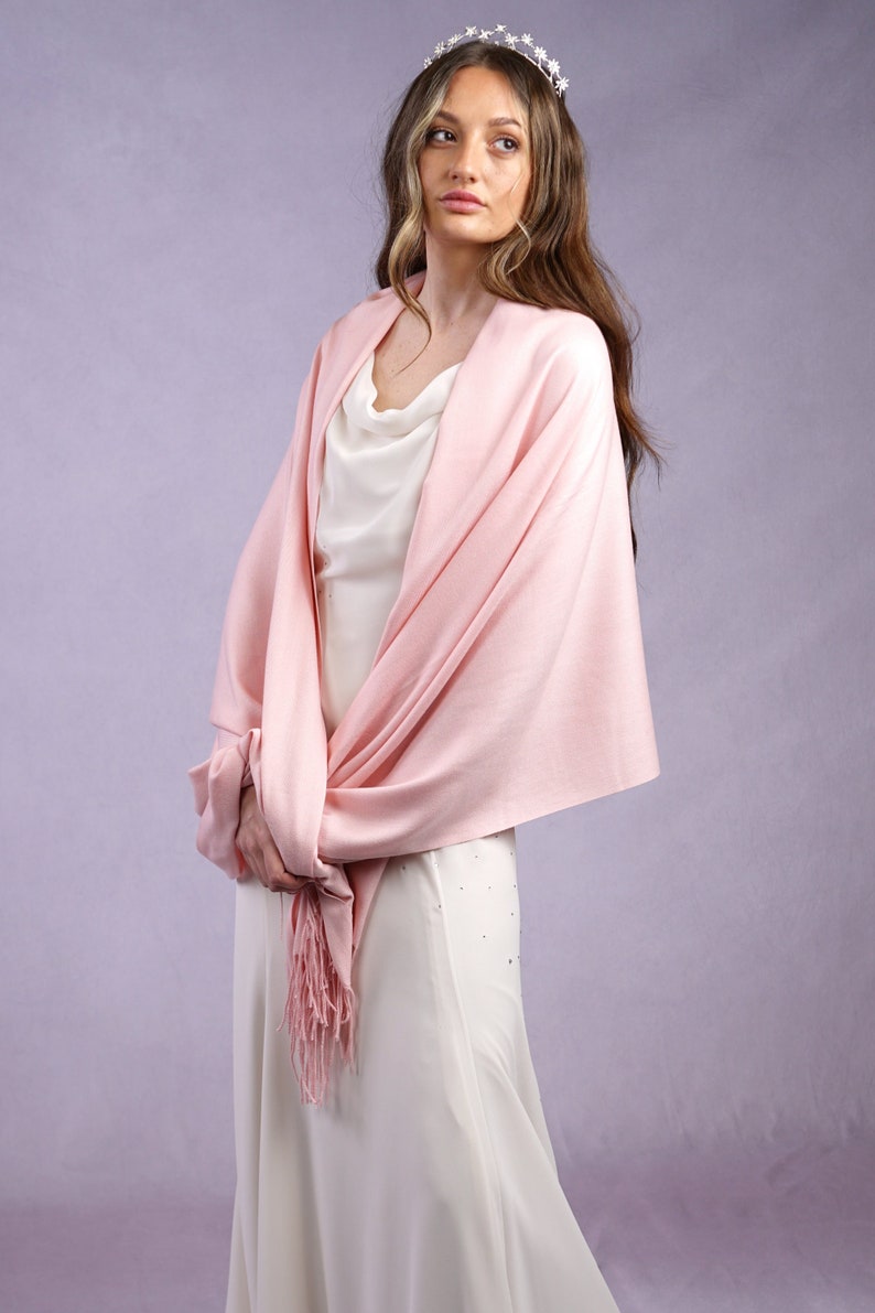 May include: A light pink shawl with a fringed edge is draped over a white dress. A silver tiara with star-shaped accents adorns the hair. The shawl is knotted at the front, creating a layered effect. The dress has a cowl neckline.