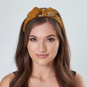May include: A woman with long brown hair is wearing a mustard yellow headband with a floral pattern. The headband is tied in a bow on top of her head.