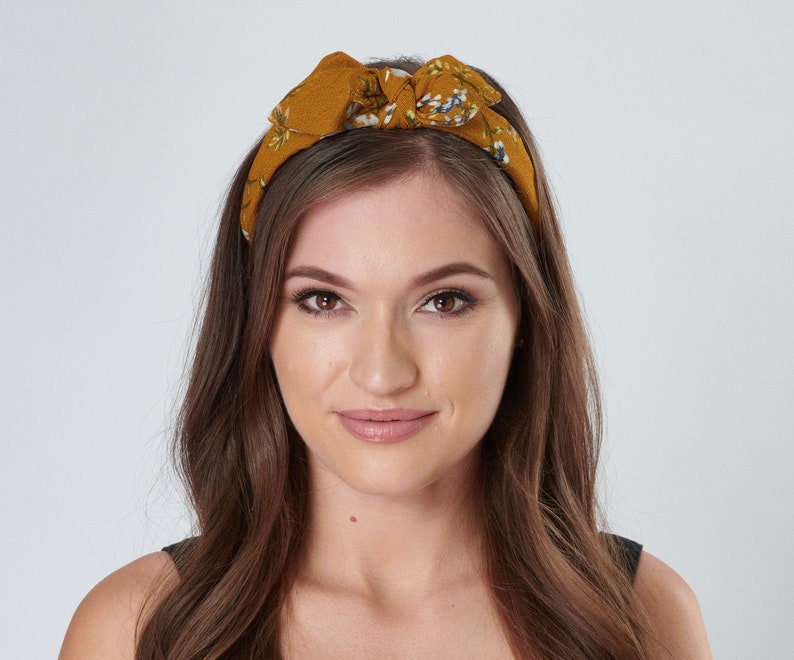 May include: A woman with long brown hair is wearing a mustard yellow headband with a floral pattern. The headband is tied in a bow on top of her head.