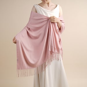 May include: A soft, light pink rectangular shawl featuring a smooth, solid-colored fabric texture and delicate fringe detailing along the edges. The wrap is draped across the shoulders and arms, showcasing its fluid, lightweight drape and length.