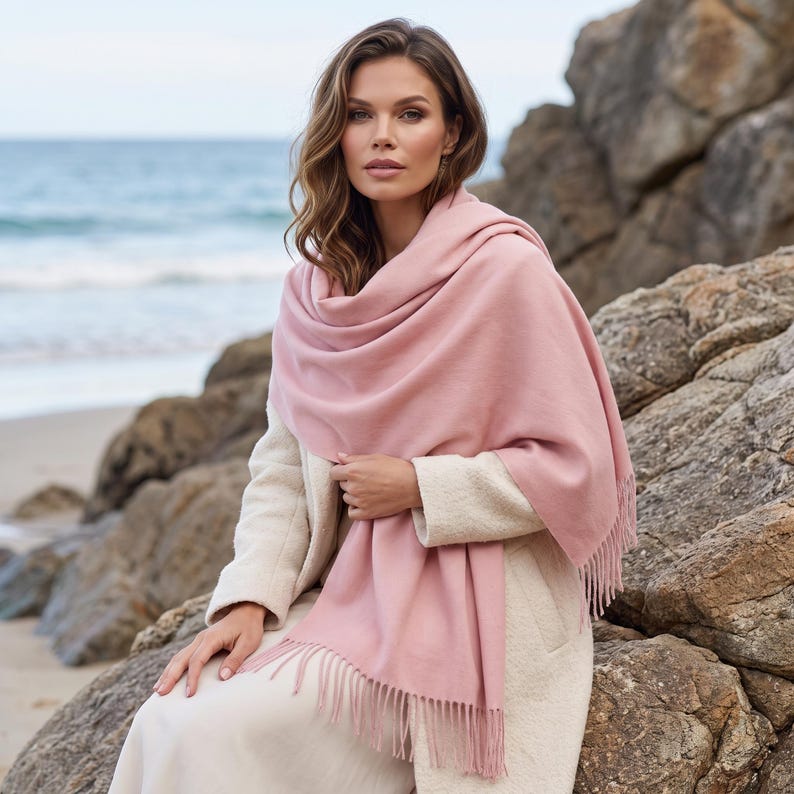 May include: A soft, solid light pink scarf with a smooth texture and fringed edges. The scarf is draped over the shoulders and upper body, featuring a rectangular shape with delicate tassels along the bottom hem, providing a simple, clean design.