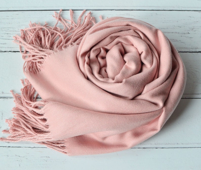 May include: A rolled-up, light pink scarf with fringed edges. The scarf is made of a soft material and is displayed on a white wooden surface. The scarf's color is a muted, dusty rose, and the fringe adds a textural element.