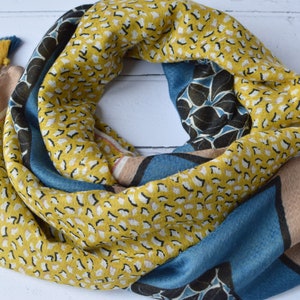 May include: Mustard yellow scarf with a repeating white and black dot pattern. Sections of teal blue and tan feature floral and geometric designs. The scarf is finished with yellow and teal blue tassels.
