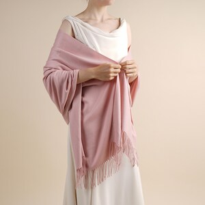 May include: A soft, solid dusty pink shawl draped over the shoulders. The rectangular wrap features a smooth, fine-knit texture and is finished with delicate, matching fringe along the short edges, creating a simple and elegant accessory.