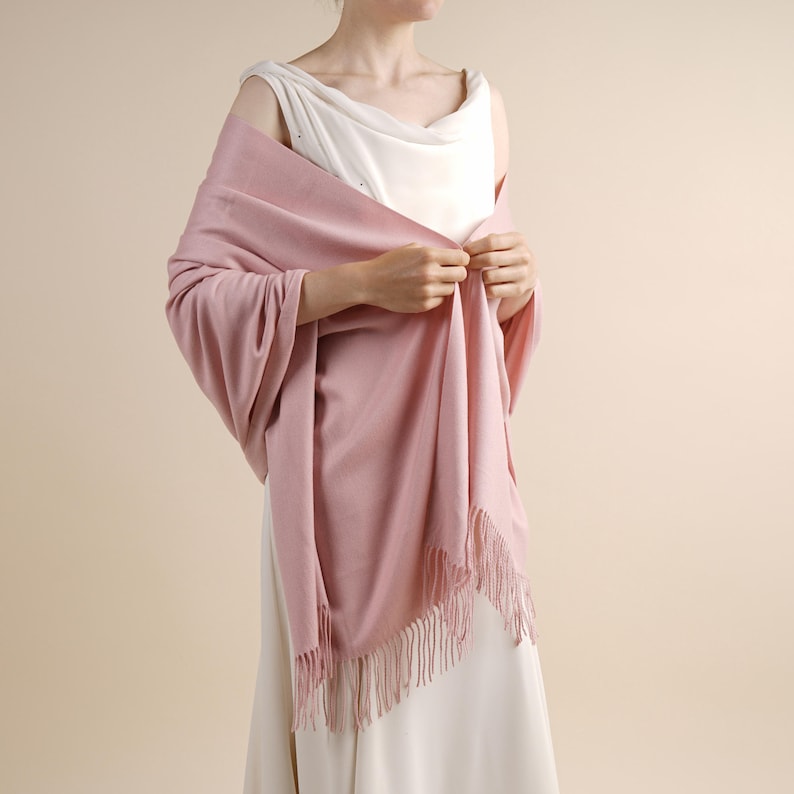 May include: A soft, solid dusty pink shawl draped over the shoulders. The rectangular wrap features a smooth, fine-knit texture and is finished with delicate, matching fringe along the short edges, creating a simple and elegant accessory.