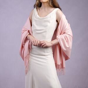 May include: A woman wearing a white sleeveless gown with a cowl neckline and a light pink shawl. The shawl has fringe. She is also wearing a silver tiara with star-shaped accents. The background is a light purple.