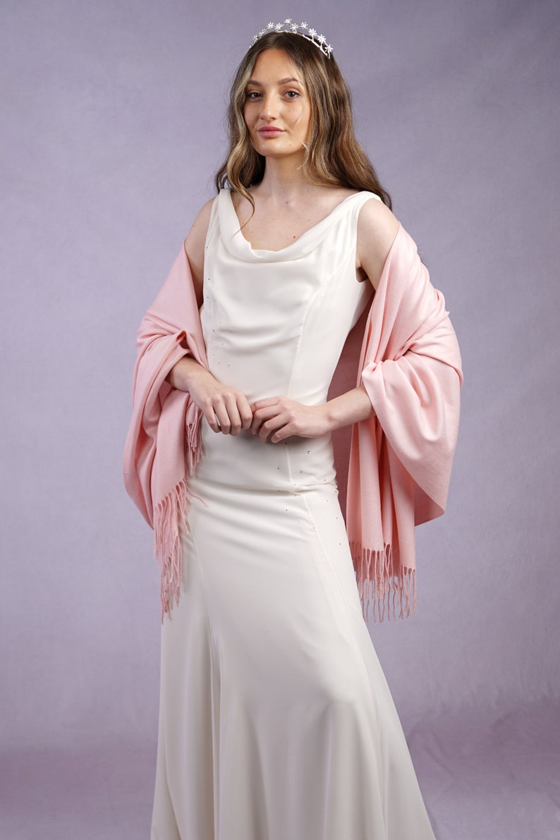 May include: A woman wearing a white sleeveless gown with a cowl neckline and a light pink shawl. The shawl has fringe. She is also wearing a silver tiara with star-shaped accents. The background is a light purple.