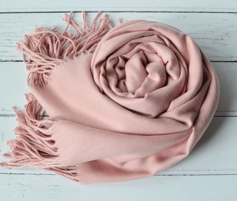 May include: A soft, blush pink scarf with a smooth, fine-knit texture and long, twisted fringe detailing at the ends. The scarf is loosely coiled into a circular shape, showcasing the consistent pastel pink color and the delicate fabric weave.