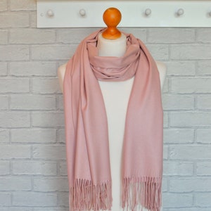 May include: A blush pink scarf with fringe detail is draped over a mannequin. The scarf is made of a soft material and is displayed against a white brick wall. The scarf is a solid color and has a simple design.