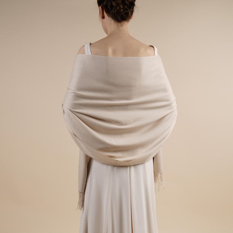 May include: A beige shawl draped over the shoulders, with a soft, flowing texture. The shawl has a subtle sheen and fringe detailing at the ends. The garment is worn over a cream-colored dress, creating a cohesive and elegant look.