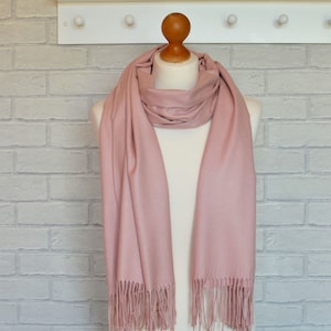 May include: A solid dusty rose scarf draped around a mannequin neck. The scarf features a smooth, matte texture and is finished with delicate, straight fringe tassels along both bottom edges, creating a simple and clean appearance for this accessory.