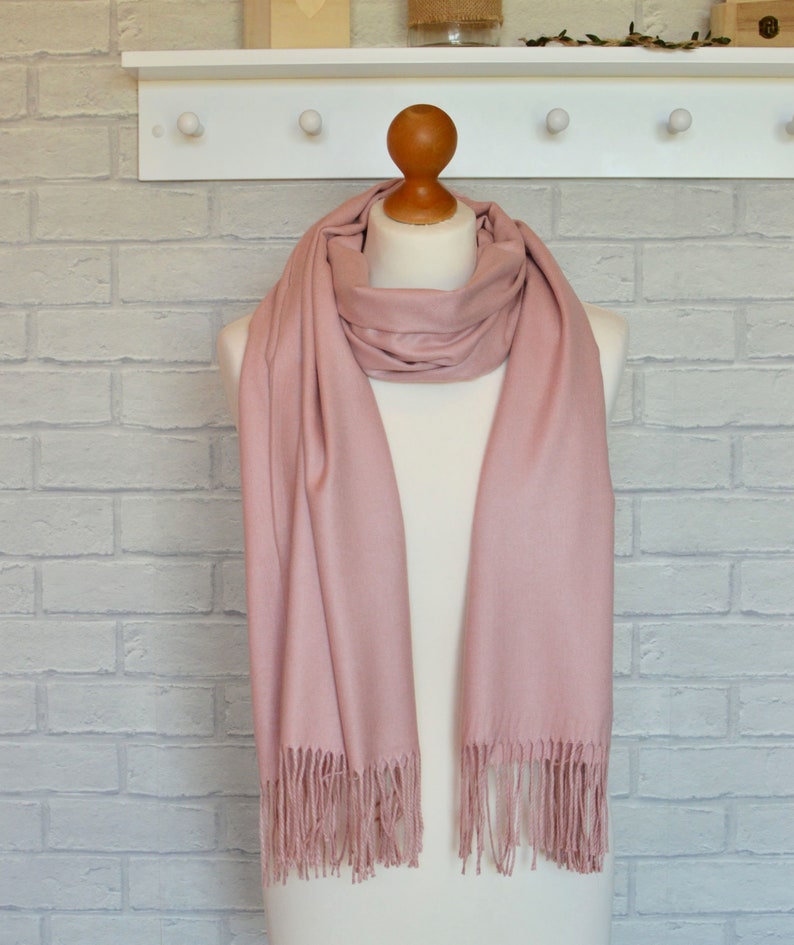 May include: A solid dusty rose scarf draped around a mannequin neck. The scarf features a smooth, matte texture and is finished with delicate, straight fringe tassels along both bottom edges, creating a simple and clean appearance for this accessory.