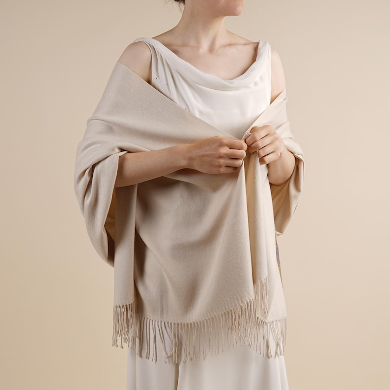 May include: A beige shawl with fringe detail. The shawl is draped over the shoulders and arms. The shawl is made of a soft material and has a simple, elegant design. The shawl is a neutral color and would complement many outfits.