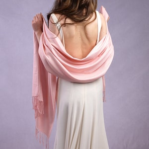 May include: A light pink shawl with fringe detail draped over a white dress. The shawl is wrapped around the shoulders. A silver tiara is worn on the head. The background is a light purple color.