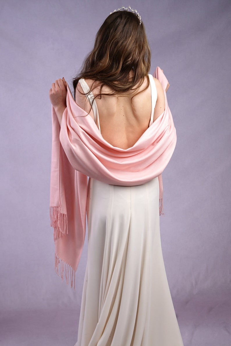 May include: A light pink shawl with fringe detail draped over a white dress. The shawl is wrapped around the shoulders. A silver tiara is worn on the head. The background is a light purple color.