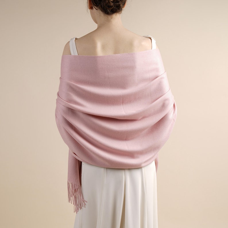 May include: A pale pink shawl draped over the shoulders. The shawl has a soft texture and a fringed edge. The person is wearing a white top and the background is a neutral colour.