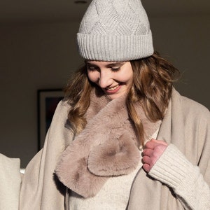 Peut inclure: Une femme portant un bonnet en tricot gris clair et une écharpe en fausse fourrure marron clair. L'écharpe est enroulée autour de son cou et nouée à l'avant. Elle sourit et regarde vers le bas.