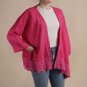May include: A bright pink linen cardigan with a delicate eyelet trim. The cardigan has a relaxed fit and a slightly cropped length.