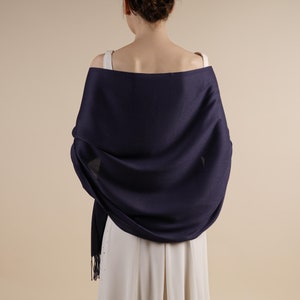 Könnte beinhalten: Ein marineblauer Schal, der über den Schultern drapiert ist. Der Schal ist aus einem weichen Stoff gefertigt und hat einen Fransenrand. Das Model trägt ein weißes Kleid. Der Hintergrund ist einfarbig beige.