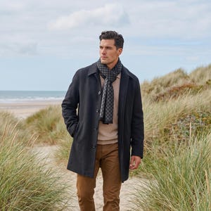 Könnte beinhalten: Ein dunkelgrauer Mantel über einem beigen Pullover und einer braunen Hose, kombiniert mit einem dunkelgrauen Schal mit wiederkehrendem geometrischem Muster. Der Mantel hat einen klassischen Kragen, lange Ärmel und eine sichtbare Knopfleiste.