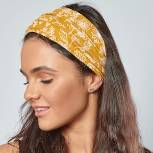Puede incluir: Una mujer con cabello largo y castaño lleva una diadema amarilla con un estampado floral blanco. La diadema está hecha de un tejido suave y elástico.