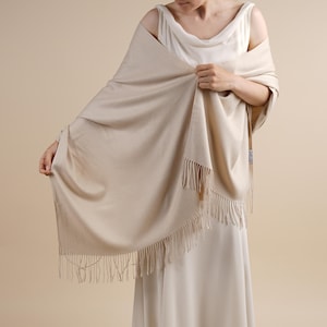 May include: A beige shawl with fringed edges. The shawl is draped over a dress. The fabric appears soft and flowing, with a subtle sheen. The neutral color makes it versatile for various outfits and occasions.