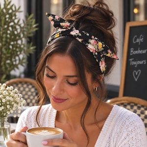 May include: A black fabric headband featuring a floral pattern with pink, yellow, and white flowers. The headband is tied in a knot at the top, creating a bow-like appearance with pointed ends. The accessory is worn in dark brown hair styled in a bun.