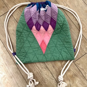 Può includere: Una borsa a cordoncino trapuntata verde, rosa, viola e blu con manici in corda bianca. La borsa ha un motivo a rombi e un piccolo cartellino che dice "Sweet Rue".