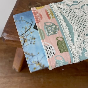 May include: A light pink fabric book cover with a white lace trim. The cover features a repeating pattern of teacups with floral and geometric designs. The cover is folded over a book with a blue and white sky and tree image.