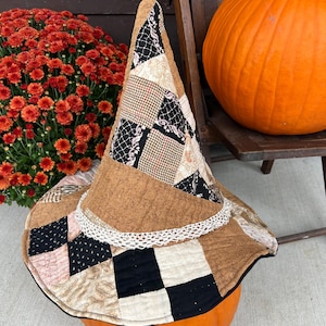 May include: A conical witch hat constructed from a patchwork of quilted fabric squares in shades of brown, black, cream, and pink, featuring a white lace trim around the base of the crown and a wide, flexible brim with visible stitching.
