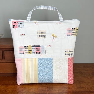 Puede incluir: Un bolso tote blanco con un estampado de mapa y edificios. La parte inferior del bolso presenta un diseño de patchwork con telas rosas, amarillas, azules y doradas. El bolso tiene cierre de cremallera y un asa con estampado de cuadros azules y blancos.