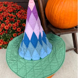 May include: A quilted pointed hat featuring a diamond pattern in shades of pink, purple, and blue, with a wide, circular green brim. The fabric has visible stitching throughout, creating a textured, padded appearance on both the cone and the brim.